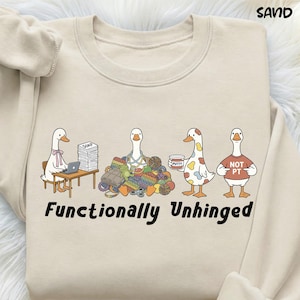 Könnte beinhalten: Sandfarbenes Sweatshirt mit einer skurrilen Illustration von vier Gänsen. Eine Gans sitzt an einem Schreibtisch mit einem Laptop, eine andere ist von Garn umgeben, eine dritte hat ein farbklecksartiges Design und die letzte trägt ein Shirt mit der Aufschrift "NOT PT". Der Text "Functionally Unhinged" steht darunter.