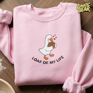Può includere: Una felpa rosa chiaro con un'oca dei cartoni animati che tiene in mano una pagnotta, con dei cuori sopra. Il testo "LOAF OF MY LIFE" è stampato sotto. La felpa ha le maniche lunghe.