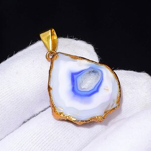 Può includere: Un ciondolo con una pietra di agata lucida con finiture e anello dorati. La pietra ha una base bianca con bande concentriche blu e grigie. Il ciondolo è tenuto su uno sfondo di tessuto bianco.