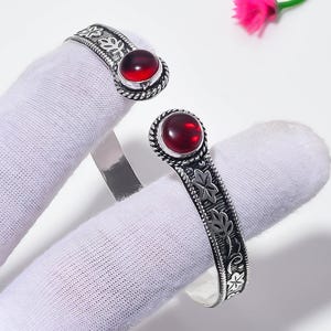 Puede incluir: Una pulsera de brazalete abierta con dos piedras cabujón redondas de color rojo intenso engastadas en cada extremo. La banda de metal plateado muestra un patrón de hojas en relieve en el exterior y un borde de cuerda trenzada alrededor.