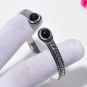 Peut inclure: Un bracelet manchette ouvert en argent oxydé, orné d'un motif de feuilles détaillé le long de l'anneau. Chaque extrémité est terminée par une pierre précieuse d'onyx noir, lisse, ronde et polie, sertie dans une bordure torsadée décorative.