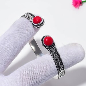 Può includere: Un bracciale a fascia aperto in metallo color argento, caratterizzato da due estremità arrotondate sormontate da pietre cabochon rosse lisce e vivaci. La fascia è decorata con un motivo a foglie in rilievo e un bordo a corda intrecciata.