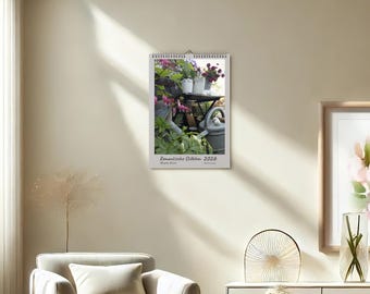 以下が含まれることがあります： 壁掛けカレンダー。庭の風景の写真が掲載されています。小さなテーブルに鉢植えの花、じょうろ、緑豊かな植物が写っています。カレンダーには「Romantic Stillleben 2026」と書かれています。