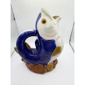May include: A ceramic pitcher in the shape of a fish. The pitcher is primarily white and blue, with brown and yellow accents. The fish has a wide open mouth and a curved tail that forms the handle.