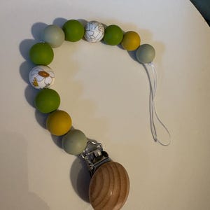 May include: A baby pacifier clip with a wooden clip and a string of colorful beads. The beads are in shades of green, yellow, and gray, with some featuring floral patterns. The clip is made of wood and metal.