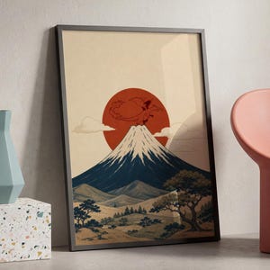 May include: A framed vertical art print featuring a stylized illustration of a snow-capped mountain peak beneath a large red sun. The landscape includes rolling hills, sparse trees, and a small dragon-like creature flying near the summit.