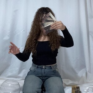May include: A person kneels, holding a deck of tarot cards in front of their face. They wear a black top and blue jeans. Several tarot cards are laid out on a white surface, along with decorative items and a glass bowl. The background is a white curtain.