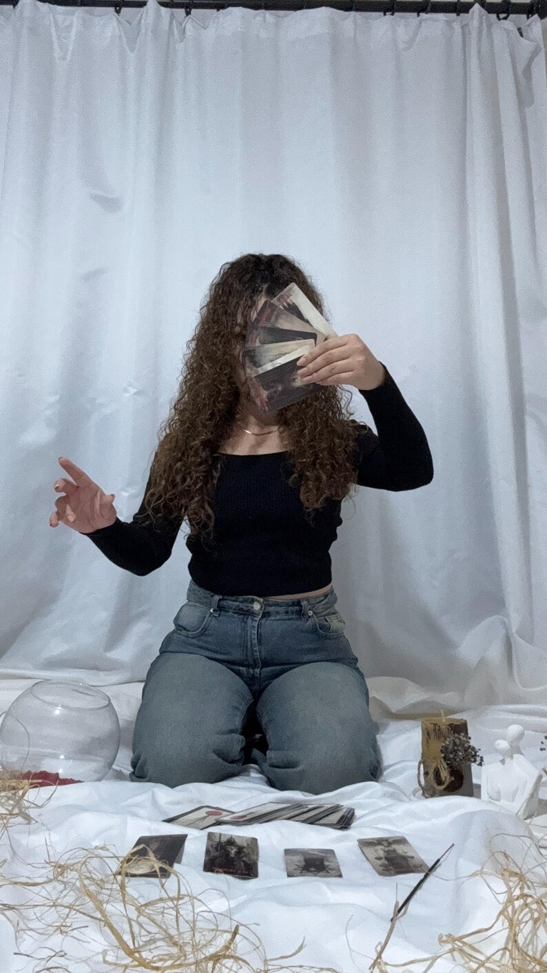May include: A person kneels, holding a deck of tarot cards in front of their face. They wear a black top and blue jeans. Several tarot cards are laid out on a white surface, along with decorative items and a glass bowl. The background is a white curtain.