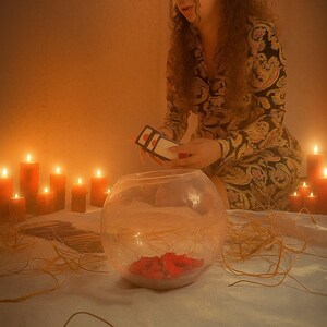 May include: A clear glass bowl with red petals inside sits on a white surface. A person holds a small rectangular object above the bowl. Lit candles and decorative straw are arranged around the bowl, creating a warm, inviting atmosphere.