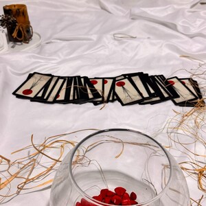 May include: A clear glass bowl filled with red stones, with a collection of cards featuring a red dot design. The cards are arranged on a white surface. A candle and dried plants are in the background.