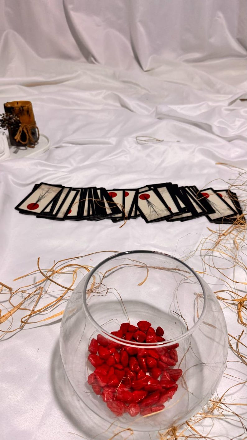 May include: A clear glass bowl filled with red stones, with a collection of cards featuring a red dot design. The cards are arranged on a white surface. A candle and dried plants are in the background.