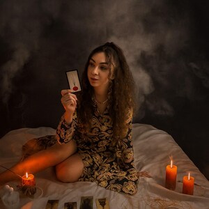 May include: A woman holding a tarot card with a red circle design, surrounded by other cards, candles, and a decorative arrangement. The woman is wearing a black and gold patterned dress. The scene is set on a white bed with a dark background.