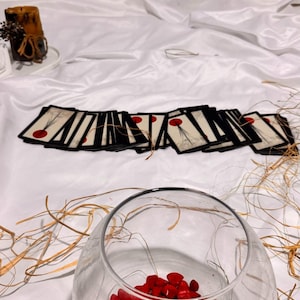 May include: A clear glass bowl filled with red stones, with a collection of cards featuring a red dot design. The cards are arranged on a white surface. A candle and dried plants are in the background.