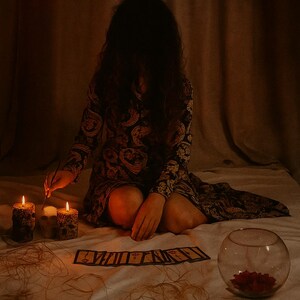 May include: A dimly lit scene featuring a person in a paisley print dress, seated on a bed with lit candles and tarot cards. A glass bowl with red flowers and twine are also present, creating a mystical atmosphere.