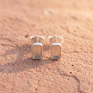 May include: A pair of silver-toned stud earrings featuring a rectangular bezel setting with a recessed center. The earrings have a smooth, metallic finish and a post back design, presented as two identical pieces side by side.