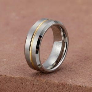 Può includere: Un anello color argento con una fascia centrale dorata. L'anello ha una finitura lucida e un bordo smussato. L'interno dell'anello è inciso con la scritta "CAHLAN TUNGSTEN".