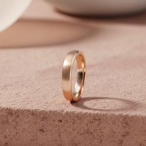 Puede incluir: Una alianza de boda de oro rosa con un acabado cepillado. El anillo está sobre una superficie rosa, con una sombra suave proyectada detrás. La banda tiene un diseño simple y clásico.