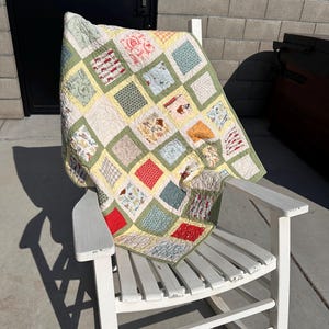 May include: A handmade quilt with a patchwork design in various colors, including red, green, yellow, and blue, is draped over a white wooden rocking chair. The quilt features square patterns with floral and geometric designs.