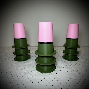 Puede incluir: Tres objetos decorativos apilados en forma de taza. Cada uno tiene una parte superior rosa, una banda negra y una base verde. Las bases están apiladas unas sobre otras, creando un diseño único. Los objetos están dispuestos sobre una superficie punteada.