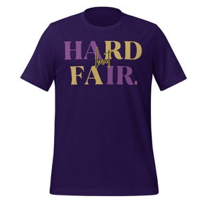 Puede incluir: Camiseta morada con la frase "HARD but FAIR" impresa. La palabra "HARD" es morada, "but" es dorada y "FAIR" es dorada y morada. Cuello redondo y mangas cortas.