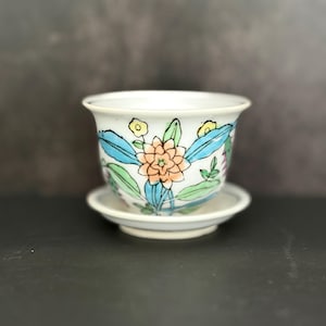 May include: White ceramic planter with a matching saucer. The planter is decorated with a floral design featuring orange, blue, and green leaves and flowers. The rim of the planter has small yellow flowers.