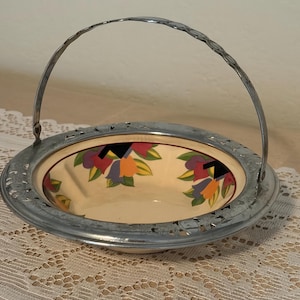 May include: A vintage serving dish with a floral design. The dish has a cream-colored interior with colorful flowers and a silver-colored metal rim and handle. The handle is arched and attached to the rim.