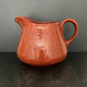May include: A terracotta-colored ceramic creamer pitcher featuring a speckled white pattern across its entire surface. The pitcher has a rounded body, a curved handle on the right side, and a small pouring spout on the upper left side.