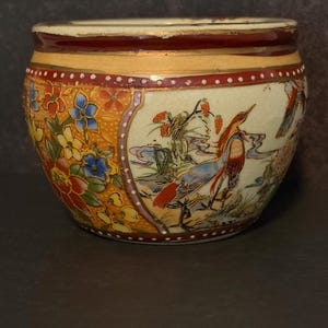 May include: A round ceramic bowl featuring a cream-colored background with hand-painted details. One side displays a vibrant floral pattern in orange, blue, and yellow, while the other side depicts a bird in a landscape. The rim is dark red with gold.