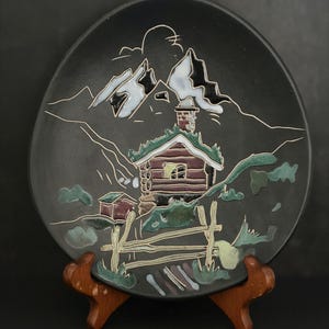 May include: A decorative black ceramic plate featuring a mountain cabin scene. The plate has a dark background with white, green, and brown accents. The cabin has a green roof and a red and brown facade. The plate is displayed on a wooden stand.