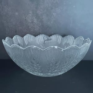 May include: A clear glass bowl with a scalloped rim. The bowl has a textured surface with a leaf-like pattern. The bowl is sitting on a dark surface against a gray background.