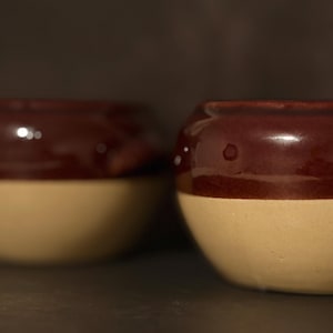May include: Two ceramic bowls with a two-tone design. The top half of each bowl is a glossy, dark brown, while the bottom half is a matte, light beige. The bowls have a rounded shape and a smooth surface.
