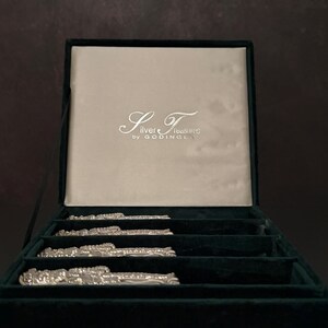 May include: A set of four silver-toned metal items with ornate, textured detailing arranged in a dark green velvet-lined presentation box. The interior lid features a light-colored satin panel with the text Silver Treasures by GODINGER.