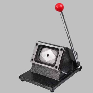 K&ouml;nnte beinhalten: Eine schwarze Metall-Stanzmaschine f&uuml;r Anstecknadeln mit strukturierter Oberfl&auml;che, ausgestattet mit einer rechteckigen Stanzform und einem langen silbernen Hebel mit rotem, kugelf&ouml;rmigem Griff f&uuml;r die manuelle Bedienung.