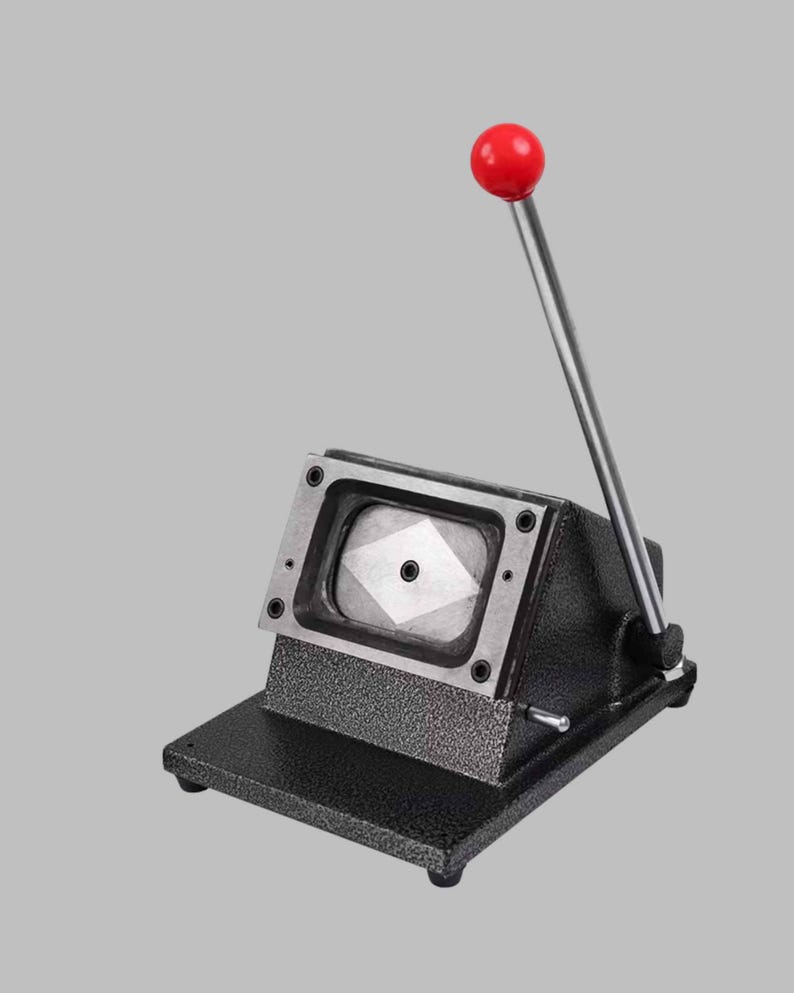 K&ouml;nnte beinhalten: Eine schwarze Metall-Stanzmaschine f&uuml;r Anstecknadeln mit strukturierter Oberfl&auml;che, ausgestattet mit einer rechteckigen Stanzform und einem langen silbernen Hebel mit rotem, kugelf&ouml;rmigem Griff f&uuml;r die manuelle Bedienung.