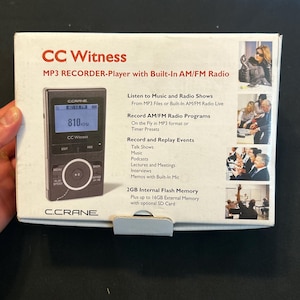 May include: A white rectangular product box for a CC Witness MP3 recorder and player with built-in AM/FM radio. The box features a product image, text detailing features like 2GB internal memory, and small photographs showing people using the device.