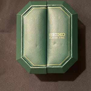 Może przedstawiać: Zielone, ośmiokątne pudełko na zegarek ze złotymi obramowaniami i napisem "SEIKO KINETIC" w złotych literach. Pudełko jest otwarte, odsłaniając wnętrze. Pudełko znajduje się na ciemnej powierzchni.