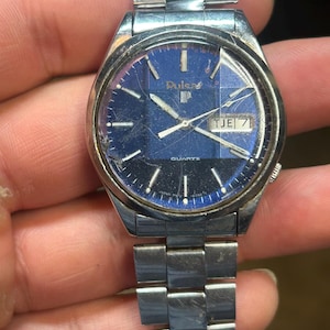May include: A vintage Pulsar quartz watch with a blue face and a silver-tone metal strap. The watch shows the day and date, with the word "Quartz" at the bottom. The watch is held in a hand.