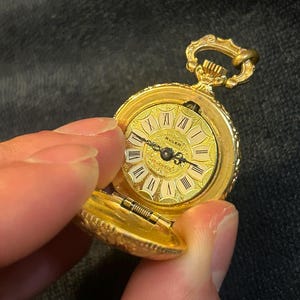 Könnte beinhalten: Eine goldene Buler-Taschenuhr mit einem detaillierten Zifferblatt mit römischen Ziffern und filigranen Zeigern. Die Uhr ist geöffnet und zeigt das komplizierte Innenleben und den Text "Buler". Die Uhr hat eine dekorative Schlaufe und einen Klappdeckel.