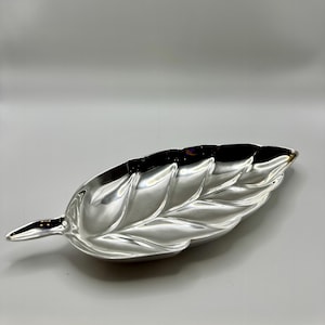 May include: A silver-colored leaf-shaped decorative tray. The tray has a detailed vein pattern and a smooth, reflective surface. The leaf design is elongated with a pointed tip and a curved shape, suitable for serving or display.