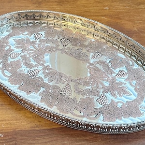 May include: An oval silver-toned metal serving tray featuring an intricate embossed pattern of grapevines, leaves, and clusters of grapes. The tray has a raised, decorative openwork gallery rim with a rope-style edge and a polished reflective centre.