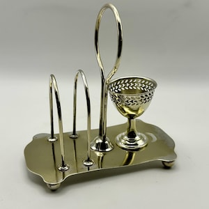 May include: Silver-toned metal egg cup and toast rack. The rack has four arched slots for toast and a decorative handle. The egg cup has a filigree design. The base is rectangular with rounded edges and small feet.