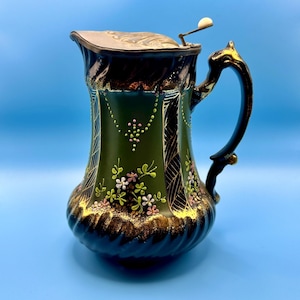May include: An antique green and black ceramic pitcher with a silver lid and handle. The pitcher is decorated with gold accents and floral patterns in pink and white. The handle has a decorative gold accent.