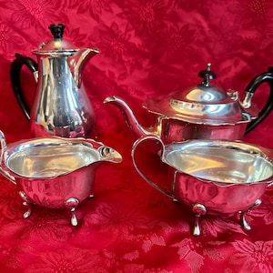 Può includere: Un servizio da tè in argento con teiera, lattiera, zuccheriera e caffettiera. La teiera e la caffettiera hanno manici e coperchi neri. Il set è su una tovaglia rossa floreale.