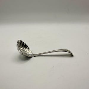 May include: A silver serving ladle with a decorative, scalloped bowl and a long, curved handle. The handle is detailed with a line of small, raised dots. The ladle is designed for serving soups, sauces, or other liquids.