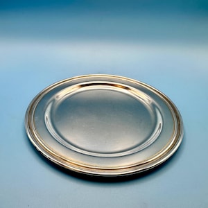 May include: A round, metallic silver-toned plate featuring a smooth, reflective center surface and a raised, polished rim. The plate has a simple, circular design with a subtle metallic sheen, showcasing a clean, unadorned aesthetic throughout.