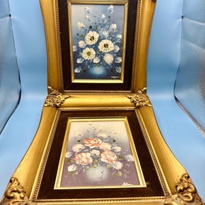 Könnte beinhalten: Zwei gerahmte Blumenbilder. Das obere Gemälde zeigt weiße Blumen mit gelben Zentren in einer blauen Vase vor blauem Hintergrund. Das untere Gemälde zeigt rosa Rosen in einer Vase. Beide Gemälde befinden sich in verzierten Goldrahmen.