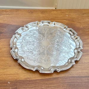 Peut inclure: Un plateau de service rond de couleur argentée présentant un motif gravé complexe de fleurs et de feuilles au centre. Le plateau possède un bord surélevé et festonné avec une finition lisse et réfléchissante, formant une bordure décorative.