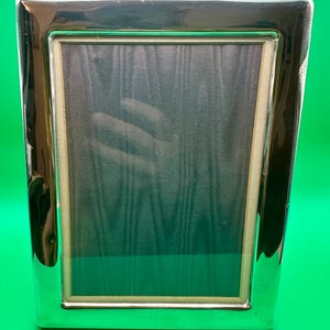May include: A rectangular picture frame with a polished, reflective silver-tone metal border. The frame features a smooth, wide metallic edge surrounding a dark, textured insert with a subtle vertical wood grain pattern and a light-coloured inner trim.