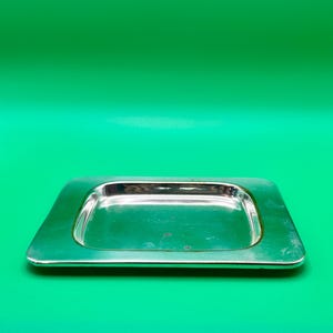 Peut inclure: Un plateau rectangulaire en métal de couleur argentée avec une surface lisse et réfléchissante et des coins arrondis. Le plateau présente une zone centrale peu profonde et encastrée entourée d'un large bord plat, offrant un aspect métallique poli.