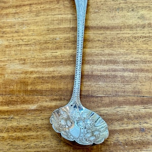 Può includere: Un cucchiaio d'argento con un ornato disegno floreale sulla coppa e sul manico. Il cucchiaio ha un bordo decorativo ed è posto su uno sfondo di legno. Il cucchiaio è lungo circa 15 cm.
